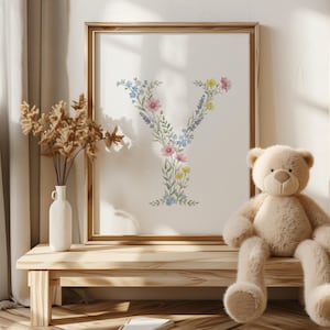 May include: A framed floral print featuring the letter "Y" composed of colorful flowers and greenery. The artwork is in a wooden frame and rests on a light-colored wooden table. A teddy bear sits nearby.