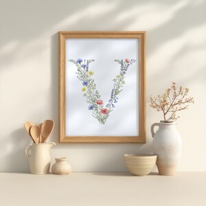May include: A framed floral art print featuring the letter "V" composed of colorful wildflowers. The wooden frame surrounds the artwork, which is displayed on a neutral-toned surface. Decorative items like a vase, bowl, and pitcher are also present.
