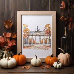 May include: A framed watercolor print of the Brandenburg Gate in Berlin, Germany, with autumn foliage. The print is displayed on a wooden surface with pumpkins and fall leaves, creating a seasonal display.