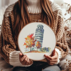 Puede incluir: Un aro de bordado de madera con una tela blanca estirada sobre él. La tela presenta un diseño de punto de cruz de la Torre Inclinada de Pisa en Italia con un árbol de colores otoñales en primer plano.