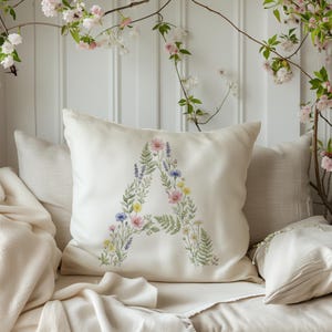 May include: A cream-colored decorative pillow with a floral "A" design. The letter is composed of various flowers in shades of pink, blue, yellow, and green. The pillow is surrounded by other pillows and a blanket, with flowering branches in the background.