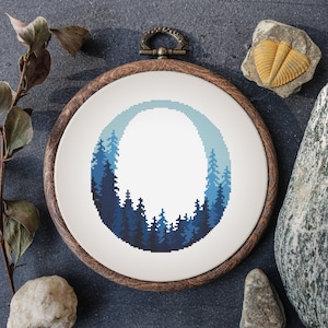 Op de afbeelding: Een borduurring met kruissteek met een boslandschap in blauwtinten. Het ontwerp is geplaatst in een houten ring met een metalen ophanglus. Het kunstwerk toont een silhouet van bomen tegen een gradiënthemel.