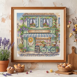 May include: A framed cross-stitch embroidery depicting a two-storey bakery with a green facade, blue striped awning, and a bicycle parked in front. The scene includes window boxes with flowers, a wreath on the door, and a menu board with text.