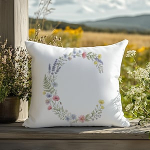 May include: White square pillow with a floral wreath design. The wreath is composed of various flowers in shades of pink, blue, yellow, and purple. The pillow is placed outdoors on a wooden surface, with a natural background.
