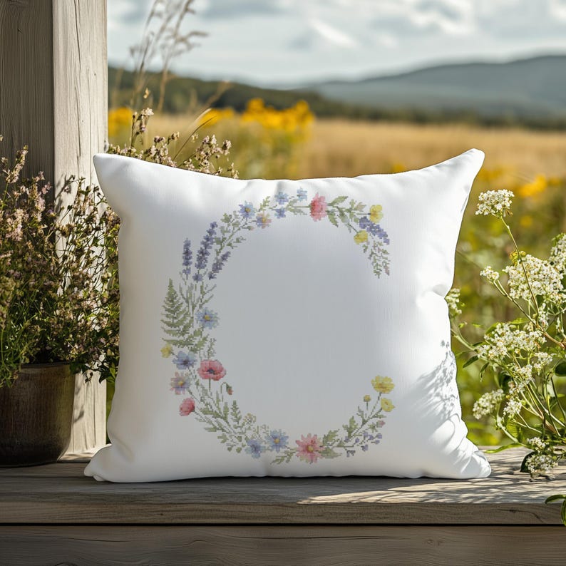 May include: White square pillow with a floral wreath design. The wreath is composed of various flowers in shades of pink, blue, yellow, and purple. The pillow is placed outdoors on a wooden surface, with a natural background.