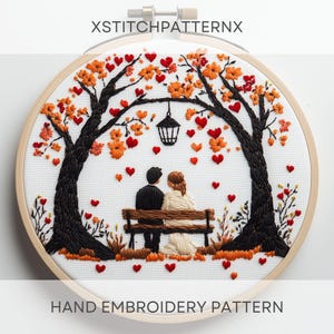 May include: Embroidered hoop art featuring a couple sitting on a bench under a tree with orange and red leaves. The couple is surrounded by small red hearts. The text "XStitchPatternx" is at the top of the hoop and "HAND EMBROIDERY PATTERN" is at the bottom.