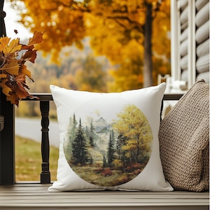May include: A white square pillow with a circular watercolor painting of a forest scene. The artwork depicts tall evergreen trees, yellow autumn trees, and a mountain range. The pillow is resting on a wooden surface.