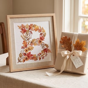 May include: A square light wood frame containing a white print of the letter B formed by autumn leaves, berries, and flowers in shades of orange, red, and yellow. Beside it sits a gift wrapped in tan paper with a cream ribbon and two dried leaves.