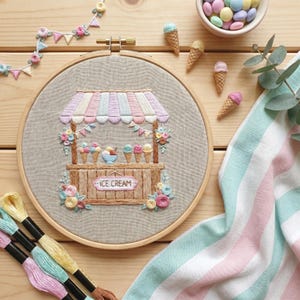 May include: An embroidered ice cream stand scene in a wooden hoop. The stand has a striped awning and is decorated with flowers. The sign reads "ICE CREAM". Colorful ice cream cones and a bowl of ice cream are featured. Pastel colors are used throughout the design.