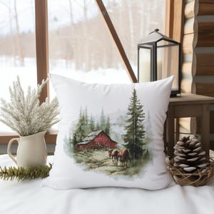 May include: White square pillow with a watercolor print of a red barn in a forest scene. Two brown horses stand near the barn. The pillow is on a white surface, with a vase of dried flowers and a pinecone nearby.