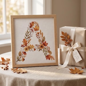May include: A framed print featuring the letter A composed of autumn leaves, berries, and flowers in shades of orange, yellow, and brown. Beside it sits a gift box wrapped in tan paper, tied with a cream ribbon, and adorned with a small leaf sprig.