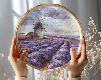 May include: A round wooden frame with a gold trim holds a cross-stitch embroidery of a lavender field with a windmill in the distance. The sky is a light blue with white clouds.