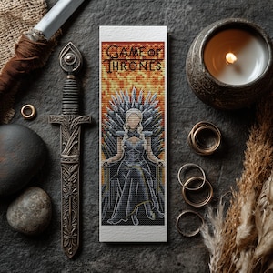 Puede incluir: Un punto de cruz marcapáginas con un diseño de Juego de Tronos. Muestra un personaje en el Trono de Hierro, con el título de la serie en la parte superior. El fondo es un degradado naranja y amarillo. El marcapáginas es rectangular y blanco.