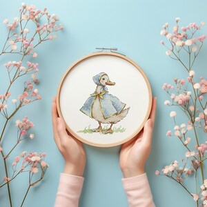 Puede incluir: Bordado de punto de cruz de un pato con capa y gorro azules, en un aro de madera. El pato est&aacute; sobre hierba verde. Flores rosas y un fondo azul claro rodean el aro.
