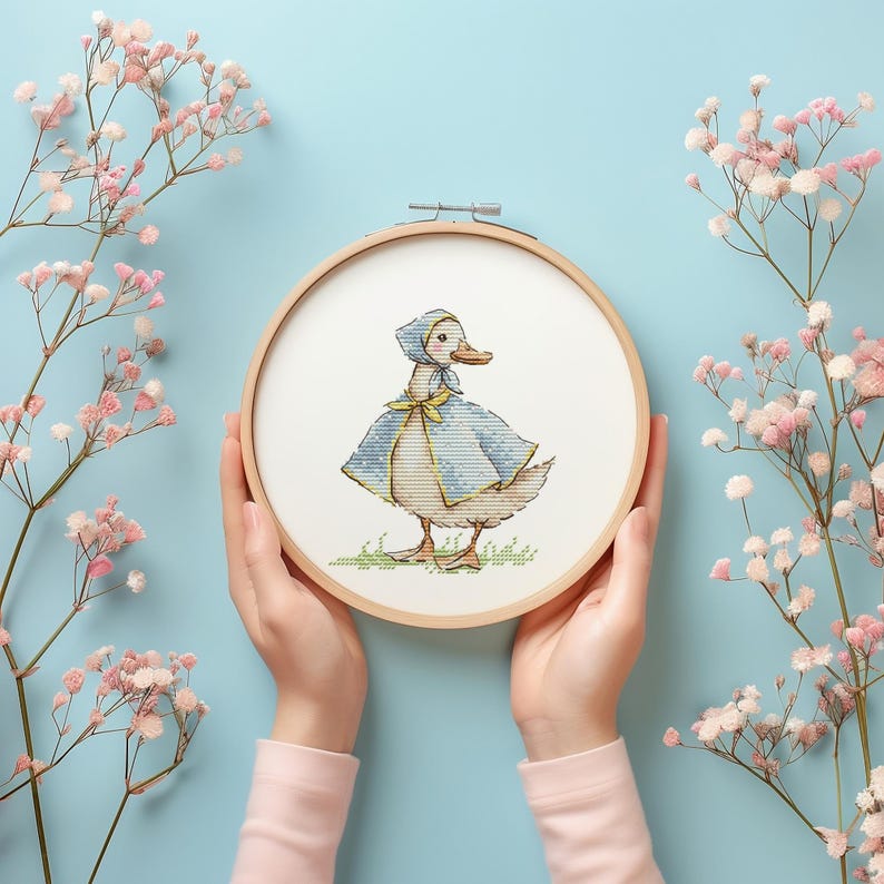 Puede incluir: Bordado de punto de cruz de un pato con capa y gorro azules, en un aro de madera. El pato est&aacute; sobre hierba verde. Flores rosas y un fondo azul claro rodean el aro.