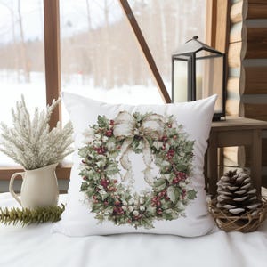 Könnte beinhalten: Ein weißes quadratisches Kissen mit einer detaillierten Illustration eines Weihnachtskranzes. Der Kranz besteht aus grünen Blättern, roten Beeren und einer großen beigen Schleife. Das Kissen liegt auf einer weißen Oberfläche, mit einem Fenster und dekorativen Gegenständen im Hintergrund.