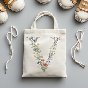 May include: A white canvas tote bag with a floral "V" design in blue, yellow, and red. The bag has two handles and is surrounded by white shoelaces and two pairs of tan and white sneakers.
