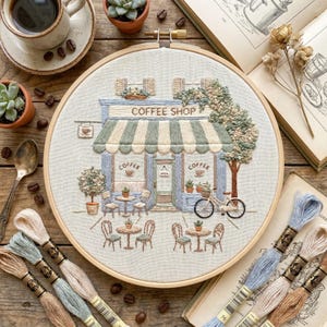 May include: Embroidered coffee shop scene in a wooden hoop. The design features a blue and green striped awning, tables, chairs, and a bicycle. The words "COFFEE SHOP" are stitched above the entrance. Coffee cups and succulents are also visible.