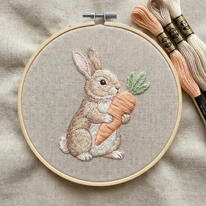 May include: A hand-stitched embroidery of a rabbit clutching a carrot, set within a wooden hoop. The rabbit is depicted in shades of brown and white, with an orange carrot and green leaves. Embroidery threads in peach and cream are visible.