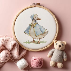 Puede incluir: Un bordado de punto de cruz de un pato con capa y gorro azules, en un aro de madera. La imagen incluye hilo rosa, una bola de hilo blanco, un osito de peluche y un fondo rosa, lo que sugiere un tema artesanal o de guarder&iacute;a.