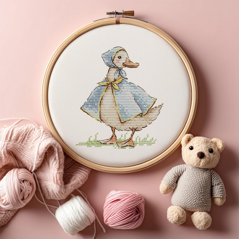 Puede incluir: Un bordado de punto de cruz de un pato con capa y gorro azules, en un aro de madera. La imagen incluye hilo rosa, una bola de hilo blanco, un osito de peluche y un fondo rosa, lo que sugiere un tema artesanal o de guarder&iacute;a.