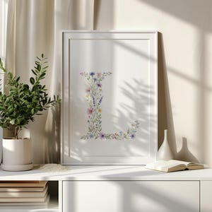 Puede incluir: Una impresión floral enmarcada con la letra "L" compuesta de coloridas flores de acuarela. La obra de arte se exhibe sobre una superficie blanca, con una planta en una maceta blanca y un libro cerca. El fondo incluye una cortina beige.