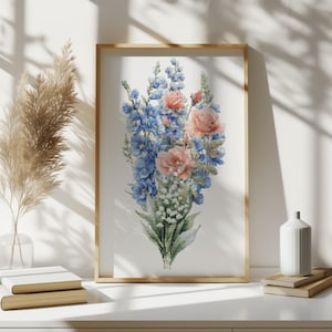 Op de afbeelding: Een ingelijst kruissteekkunstwerk met een bloemboeket van blauwe riddersporen, perzikrozen en witte lelietjes-van-dalen. Het kunstwerk staat tegen een witte achtergrond, is ingelijst in een licht houten frame en wordt tentoongesteld op een wit oppervlak.