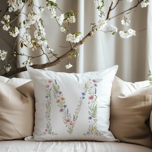Puede incluir: Cojín decorativo blanco con un diseño floral de la letra "M". La letra está compuesta por flores y vegetación coloridas, incluyendo amapolas rosas, acianos azules y lavanda. El cojín está sobre un sofá beige, con una rama floreciente en el fondo.