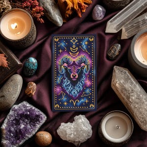 May include: An embroidered Aries zodiac sign card with a navy blue background, gold trim, and a ram's head design in purple, blue, and yellow. The card is surrounded by crystals, candles, and other spiritual items on a dark purple satin surface.