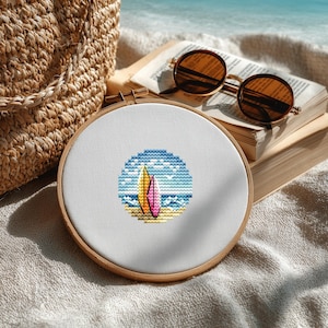 Puede incluir: Un bastidor de bordado de punto de cruz con una escena de playa con dos tablas de surf. El diseño incluye una tabla de surf amarilla y rosa, el océano azul y una playa de arena. El bastidor se encuentra sobre un fondo de una bolsa tejida, gafas de sol y un libro.