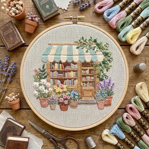 May include: Embroidered hoop art featuring a bookstore scene with a striped awning, bookshelves, and potted flowers. The embroidery includes various colors of thread, with miniature books and embroidery floss spools surrounding the hoop. A thimble and scissors are also present.