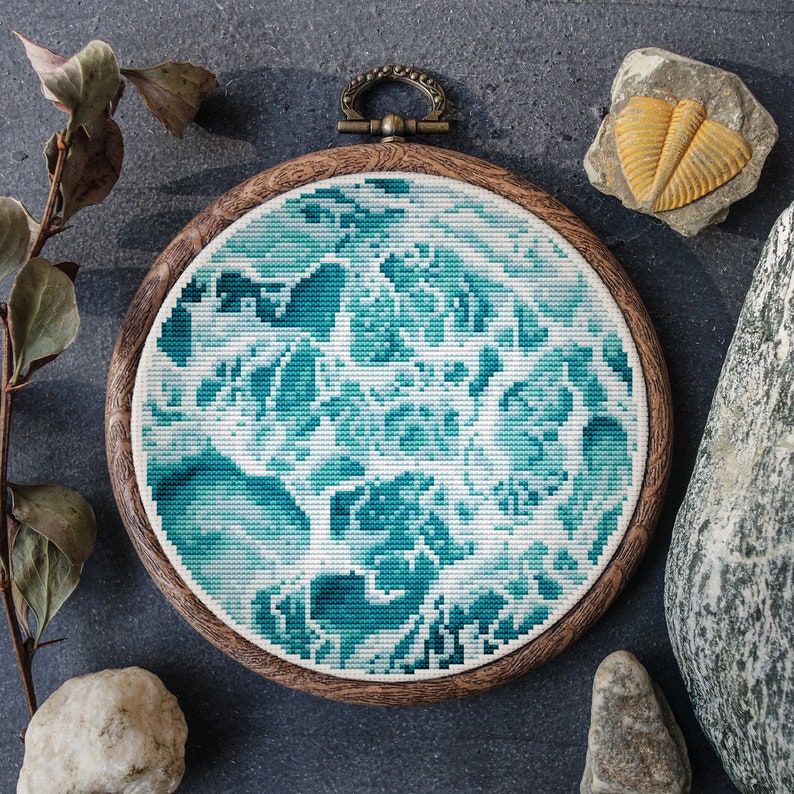 Pu&ograve; includere: Un cerchio da ricamo rotondo in legno con un disegno a punto croce blu e bianco a forma di vortice. Il cerchio &egrave; su una superficie grigia testurizzata.