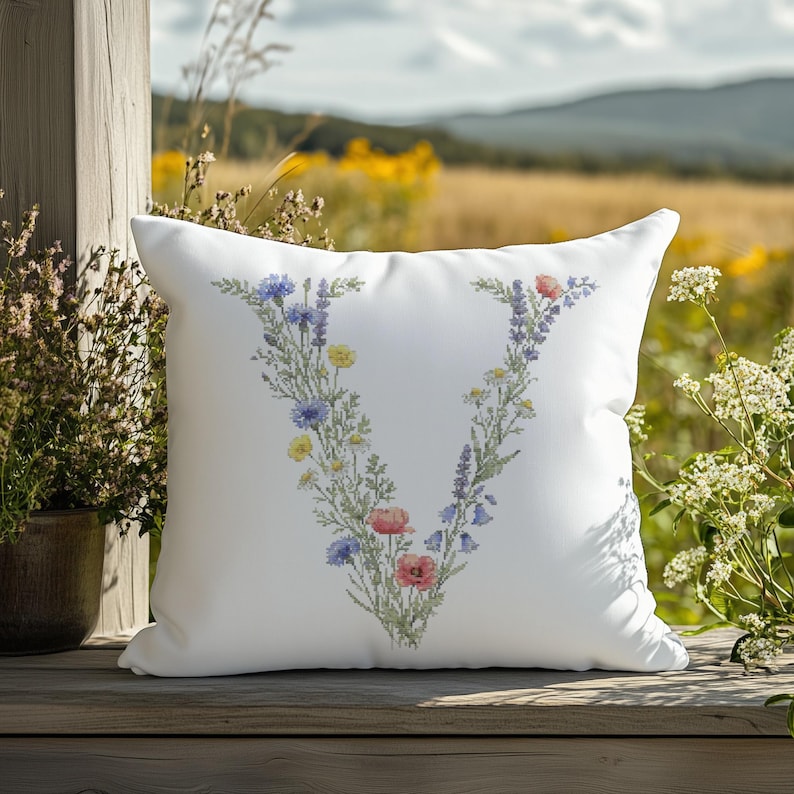 May include: White square pillow with a floral "V" design. The embroidered letter is filled with colorful flowers, including blue cornflowers, pink poppies, and yellow blooms. The pillow is placed on a wooden surface with a natural outdoor background.