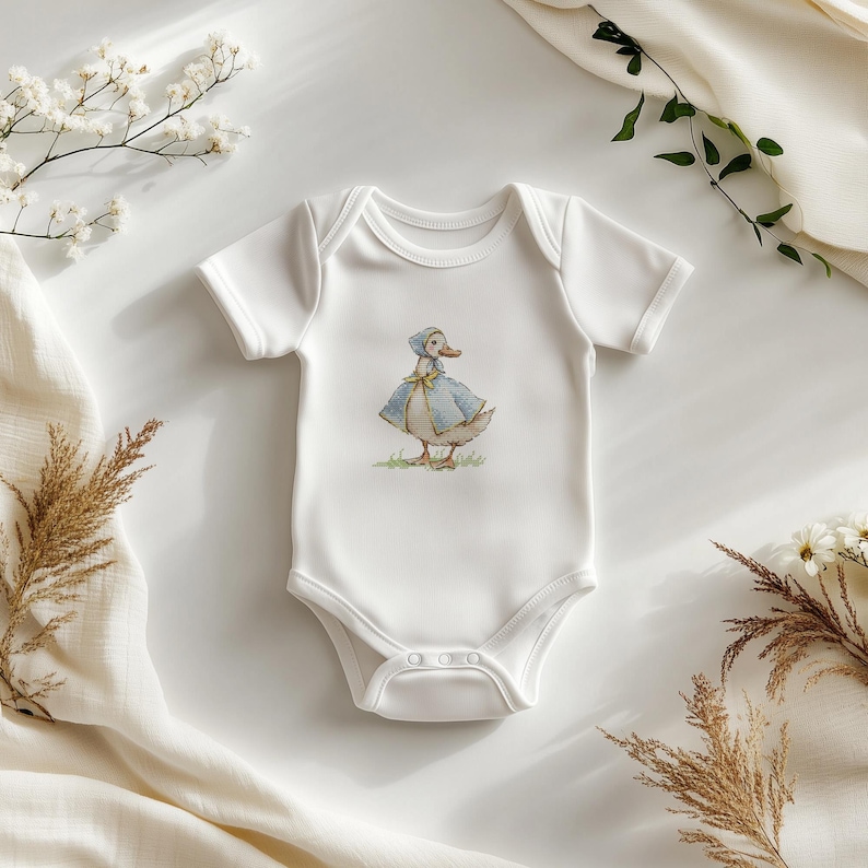 Puede incluir: Body de beb&eacute; blanco con una ilustraci&oacute;n en acuarela de un pato con capa y gorro azules. El pato est&aacute; sobre hierba verde. El body tiene mangas cortas y cierres de presi&oacute;n.