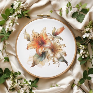 Puede incluir: Primer plano de un aro de bordado de punto de cruz con un ramo de lirios naranja y blanco con hojas verdes sobre un fondo blanco.