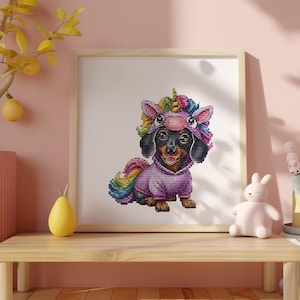May include: A framed cross-stitch of a dachshund wearing a unicorn costume. The dog is black and brown, with a pink hoodie and a colorful mane and tail. The artwork is displayed on a wooden table with decorative items.