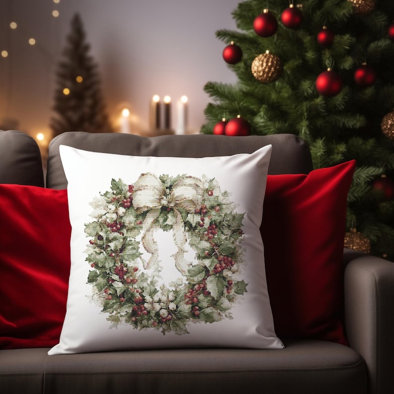 May include: A white square pillow with a watercolor design of a Christmas wreath. The wreath features green leaves, red berries, and a cream-colored bow. The pillow is on a gray couch with a red pillow and a Christmas tree in the background.