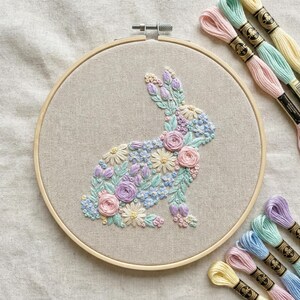 Puede incluir: Arte de aro de conejo bordado con un diseño floral en colores pastel. El conejo está lleno de flores rosas, azules y moradas, sobre una tela beige. Hilos de bordar en colores a juego están dispuestos cerca.