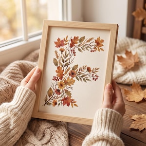 Op de afbeelding: Een lichthouten lijst met een kruissteekborduurwerk van de letter F, gevormd door een botanisch arrangement van herfstbladeren, bessen en kleine bloemen in tinten oranje, rood, geel en bruin op een crèmekleurige stoffen achtergrond.