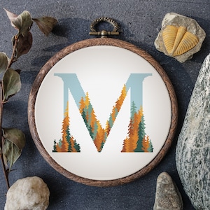 Könnte beinhalten: Ein Kreuzstich-Stickrahmen mit dem Buchstaben "M", gefüllt mit einer Waldszene in Grün-, Orange- und Blautönen. Der Holzrahmen liegt auf einer dunkelgrauen Oberfläche, umgeben von natürlichen Elementen.