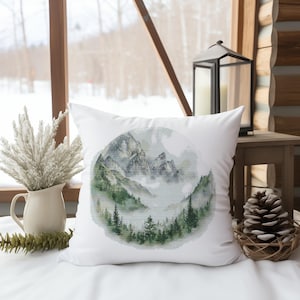 May include: White square cushion featuring a watercolour mountain scene. The design shows grey and green mountains, trees, and fog within a circular frame. The cushion is on a white surface, with a vase of dried flowers and a pinecone nearby.