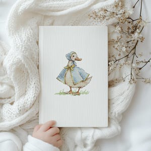 Puede incluir: Una portada de libro blanca con una ilustraci&oacute;n de punto de cruz de un pato con capa y gorro azules con un lazo amarillo. El pato est&aacute; sobre hierba verde. El libro est&aacute; sostenido por una persona.