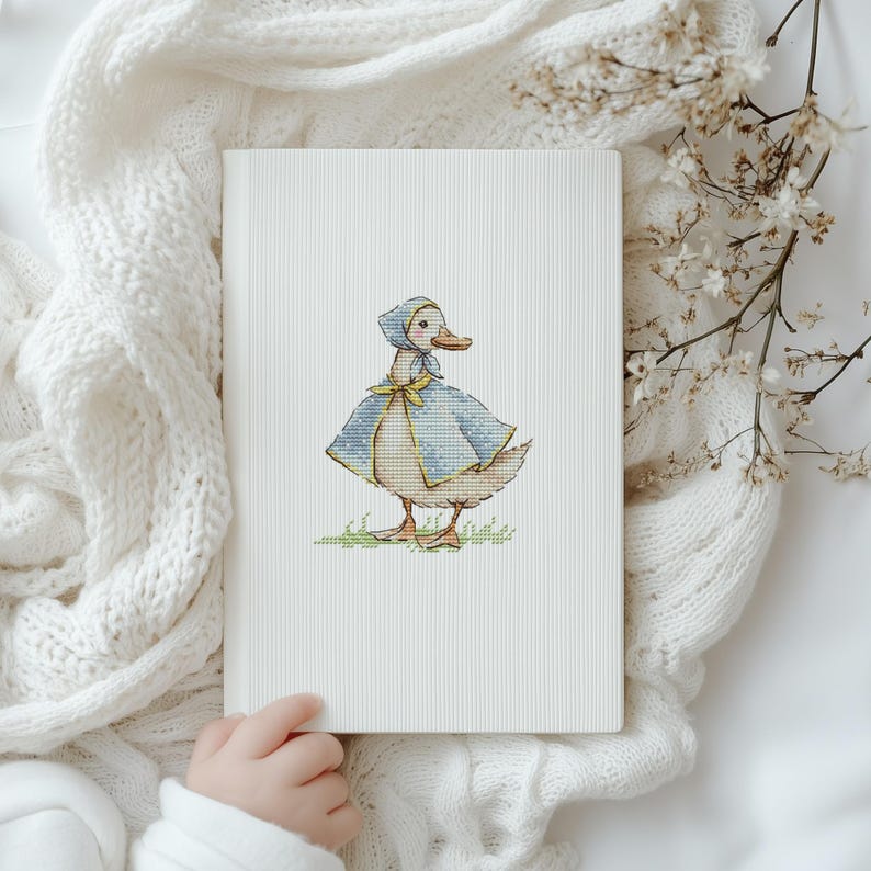 Puede incluir: Una portada de libro blanca con una ilustraci&oacute;n de punto de cruz de un pato con capa y gorro azules con un lazo amarillo. El pato est&aacute; sobre hierba verde. El libro est&aacute; sostenido por una persona.