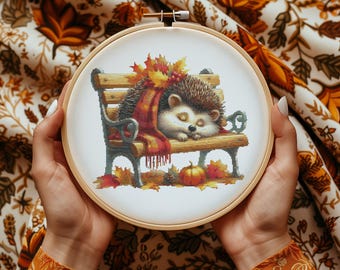 Könnte beinhalten: Ein Kreuzstich-Stickrahmen mit weißem Hintergrund, auf dem ein braunes und weißes Igel auf einer Holzbank mit einem rot-braunen Schal schläft. Die Bank ist mit Herbstblättern und einem kleinen orangefarbenen Kürbis verziert.