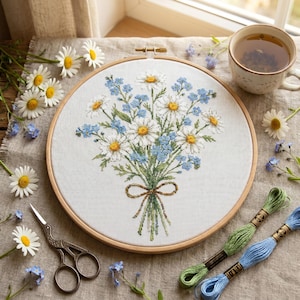 May include: A circular wooden embroidery hoop containing a white fabric panel with a cross-stitched bouquet of white daisies with yellow centers and small blue flowers, tied with a brown thread bow, accompanied by embroidery scissors and thread.