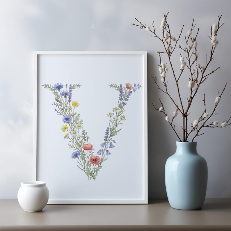 May include: A framed print featuring the letter "V" composed of colorful wildflowers. The floral arrangement includes blue cornflowers, yellow blooms, red poppies, and lavender. A small white vase and a light blue vase with branches are also in the image.
