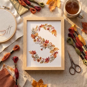 May include: A framed cross-stitch embroidery of the letter S, decorated with autumn leaves, berries, and flowers in shades of orange, red, and yellow. The piece is surrounded by embroidery hoops, colourful thread skeins, scissors, and a mug.