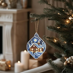 Könnte beinhalten: Ein weißer, rautenförmiger Weihnachtsbaumschmuck mit einer blauen und weißen Winterszene. Der Anhänger zeigt ein kleines braunes Haus mit einem beleuchteten Fenster und Rauch aus dem Schornstein. Das Haus ist von Bäumen und Schnee umgeben.