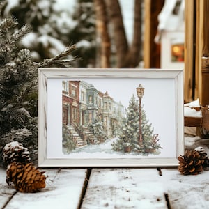 Puede incluir: Una impresión enmarcada de una escena de calle invernal con casas cubiertas de nieve y una farola. La obra de arte está en un marco blanco envejecido, sobre una superficie de madera cubierta de nieve. Piñas y ramas de pino completan el tema invernal.