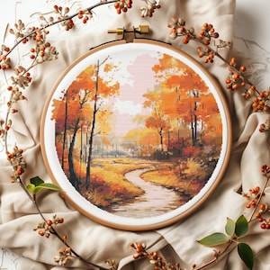 May include: A cross-stitch embroidery of a fall landscape with a winding river flowing through a forest of orange, yellow, and red trees. The embroidery is framed in a round wooden hoop.